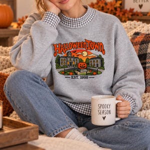 May include: A heather gray sweatshirt with a mock neck featuring a black and white gingham collar and cuffs. The front displays a graphic of a building, a large orange pumpkin, and the text HALLOWEENTOWN EST. 1998. A white mug reads SPOOKY SEASON.