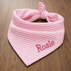 May include: A pink and white seersucker pet bandana featuring a triangular shape with a tied knot at the top. The fabric has a fine horizontal striped pattern throughout. The name Rosie is embroidered in a dark pink script font on the front.