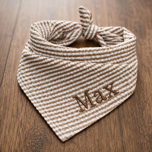May include: A triangular pet bandana featuring a brown and white seersucker striped pattern. The fabric is tied in a knot at the top. The name Max is embroidered in brown thread in a serif font on the lower front portion of the bandana fabric.