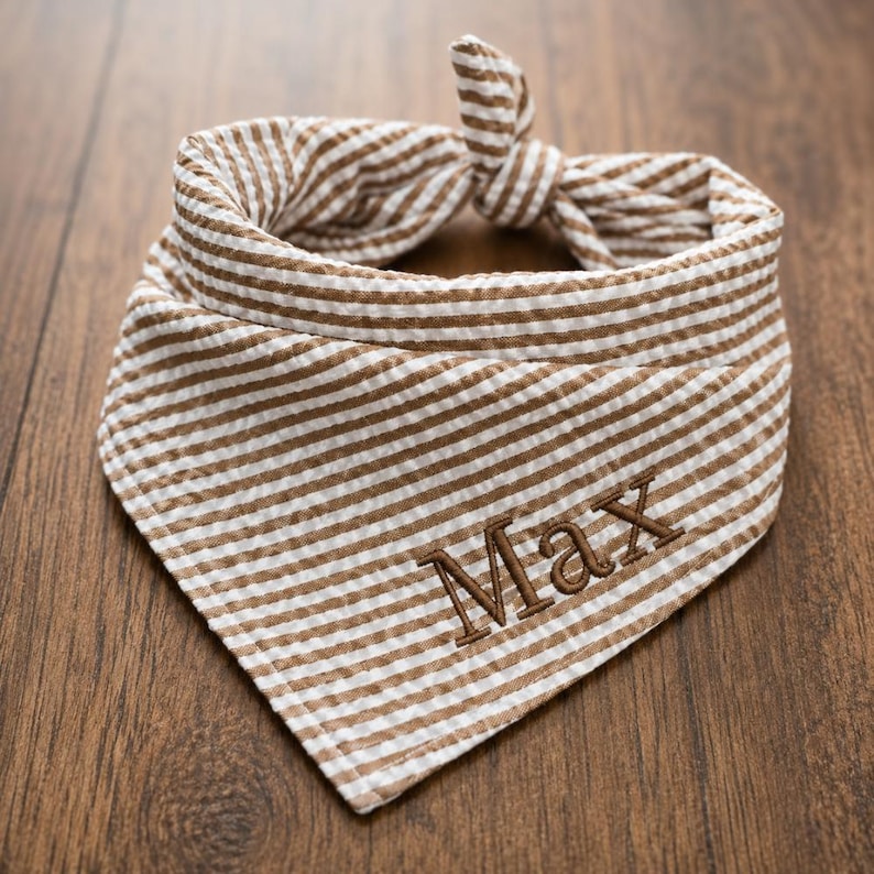 May include: A triangular pet bandana featuring a brown and white seersucker striped pattern. The fabric is tied in a knot at the top. The name Max is embroidered in brown thread in a serif font on the lower front portion of the bandana fabric.