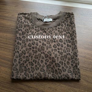 May include: Folded brown t-shirt with a leopard print pattern. The shirt has the words "custom text" printed on the front in white. A tag that says "Arlish B" is attached to the collar.