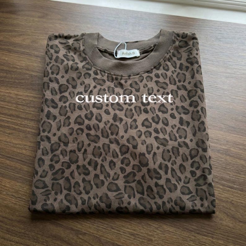 May include: Folded brown t-shirt with a leopard print pattern. The shirt has the words "custom text" printed on the front in white. A tag that says "Arlish B" is attached to the collar.