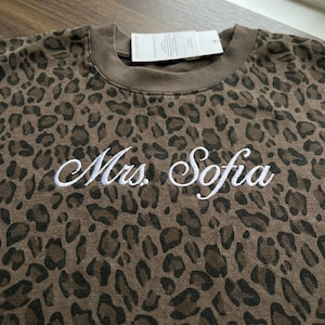 May include: Brown leopard print sweatshirt with the embroidered text "Mrs. Sofia" in white. The sweatshirt has a crew neck and a tag attached at the top. The fabric has a pattern of dark brown spots on a lighter brown background.