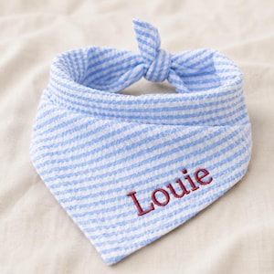 May include: A light blue and white seersucker fabric pet bandana featuring a triangular shape with a tied knot at the top. The fabric has a fine striped pattern, and the name Louie is embroidered in dark red thread on the front surface.