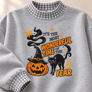 May include: A heather gray sweatshirt with a black and white gingham collar and cuffs. The front features an embroidered design of a jack-o'-lantern wearing a witch hat, a black cat, and the text IT'S THE MOST WONDERFUL TIME OF THE YEAR.