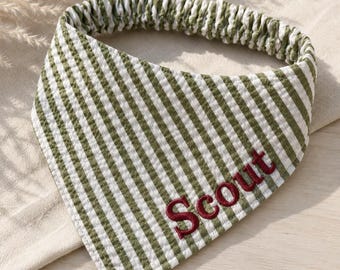 May include: A triangular pet bandana featuring a green and white seersucker striped pattern with an elasticized neck band. The word Scout is embroidered in dark red thread in a cursive font across the lower right section of the fabric.