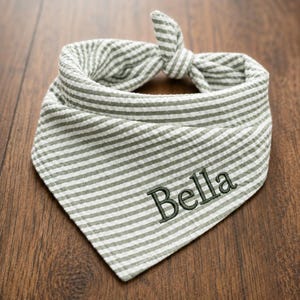 May include: A sage green and white striped pet bandana featuring a textured seersucker fabric. The bandana is tied in a knot at the top and includes the name Bella embroidered in dark green thread on the front surface of the fabric.