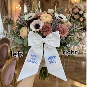 Könnte beinhalten: Ein Hochzeitsstrauß mit rosa, pfirsichfarbenen und weißen Blumen und grünem Laub. Die Stiele sind mit einer großen weißen Schleife gebunden, auf der der Text "Jose and Carol" und "HOCO 2026" in Blau gestickt ist. Blumenarrangement.