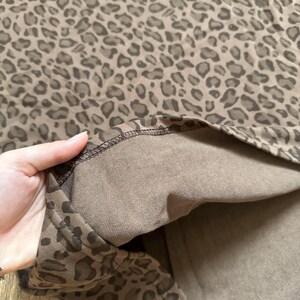 May include: A close-up of a brown leopard print fabric. The fabric has a repeating pattern of dark brown spots on a lighter brown background. The inside of the fabric is a solid brown color. The edge of the fabric is visible.