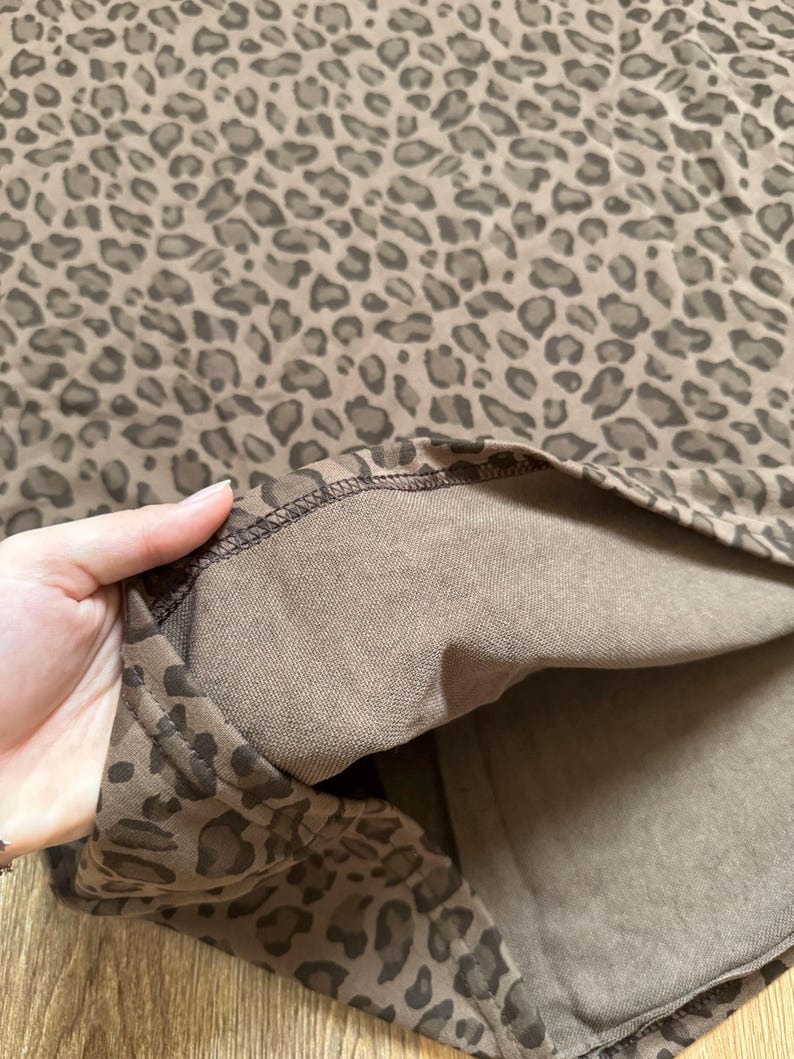 May include: A close-up of a brown leopard print fabric. The fabric has a repeating pattern of dark brown spots on a lighter brown background. The inside of the fabric is a solid brown color. The edge of the fabric is visible.
