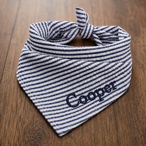 May include: A triangular pet bandana made of blue and white seersucker fabric with a striped pattern. The fabric is tied in a knot at the top. The name Cooper is embroidered in dark blue thread on the lower right side of the bandana fabric.