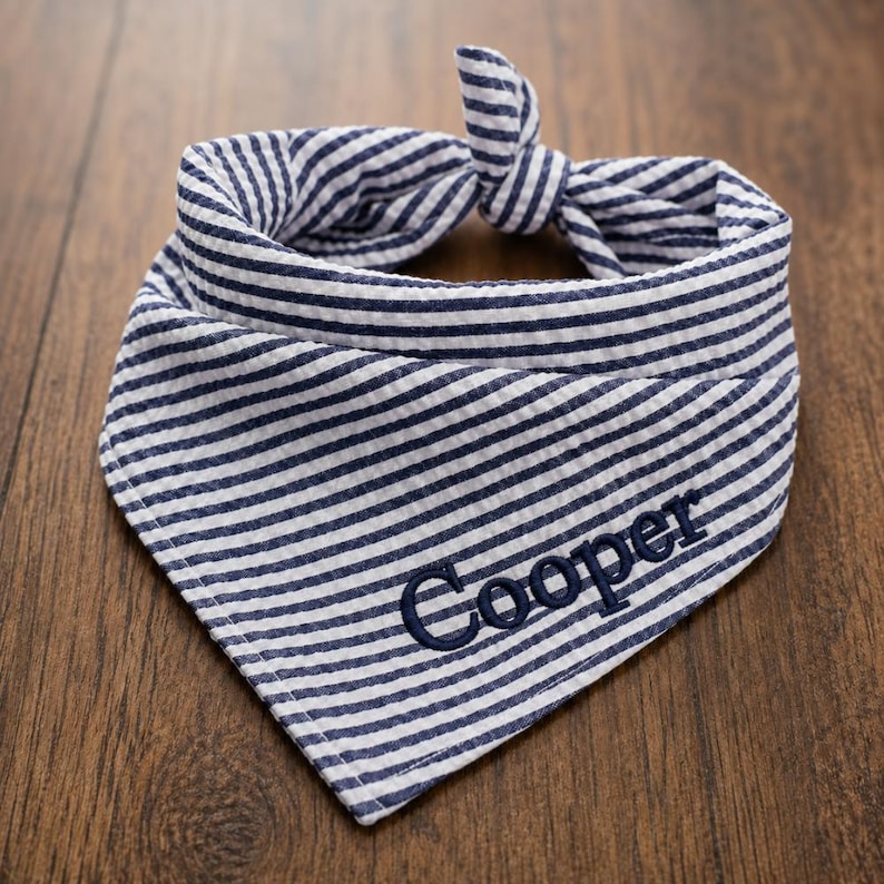 May include: A triangular pet bandana made of blue and white seersucker fabric with a striped pattern. The fabric is tied in a knot at the top. The name Cooper is embroidered in dark blue thread on the lower right side of the bandana fabric.