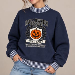 May include: A navy blue sweatshirt with a mock neck and cuffs featuring a blue and white gingham pattern. The front graphic displays a jack-o'-lantern inside a shield, with the text HALLOWEEN TOWN, EST. 1998, and WHERE BEING NORMAL IS VASTLY OVERRATED.
