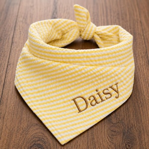 May include: A triangular pet bandana featuring a yellow and white striped seersucker pattern. The fabric is tied at the top into a knot. The name Daisy is embroidered in brown thread on the lower right side of the front of the bandana.