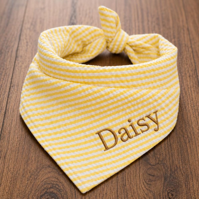 May include: A triangular pet bandana featuring a yellow and white striped seersucker pattern. The fabric is tied at the top into a knot. The name Daisy is embroidered in brown thread on the lower right side of the front of the bandana.