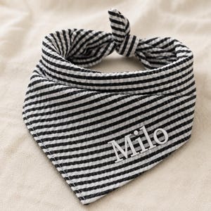 May include: A black and white striped seersucker pet bandana featuring a triangular shape with a tie-top closure. The fabric has a textured, puckered surface and the name Milo is embroidered in white capital and lowercase letters on the front.
