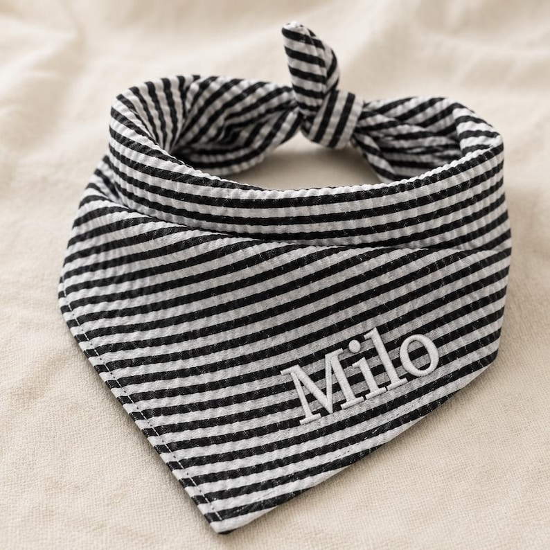 May include: A black and white striped seersucker pet bandana featuring a triangular shape with a tie-top closure. The fabric has a textured, puckered surface and the name Milo is embroidered in white capital and lowercase letters on the front.