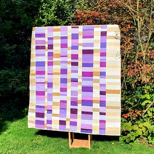 Könnte beinhalten: Eine lila und cremefarbene Patchwork-Decke mit geometrischem Muster. Die Decke wird auf einem Holzständer vor einem grünen Busch ausgestellt.