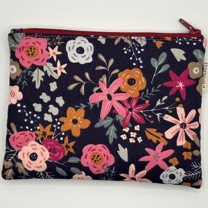 Könnte beinhalten: Eine rechteckige Tasche mit floralem Muster auf dunkelblauem Hintergrund. Das Design umfasst rosa, orange und weiße Blumen mit grünen und grauen Blättern. Sie hat einen weinroten Reißverschluss und ein kleines weißes Etikett an der Seite.