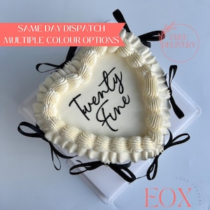May include: A heart-shaped cake with smooth off-white frosting, decorated with a piped ruffled border along the edge. The centre features the black cursive text Twenty Fine. The cake is accented with several thin black ribbons tied around the base.