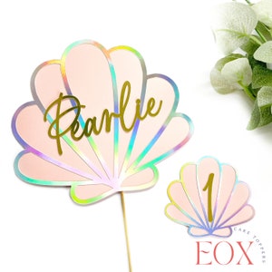 May include: Pink iridescent seashell cake topper with the name Pearlie in gold glitter lettering. A smaller seashell topper with the number 1 in gold glitter lettering is also included.