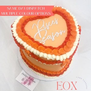 May include: A heart-shaped cake decorated with orange and white frosting. The cake is topped with a white frosting design that says "Pisces Season".