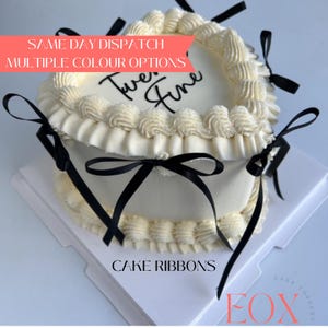 May include: A heart-shaped cake with smooth white frosting, decorated with piped cream borders along the top and bottom edges. The cake is adorned with several thin black ribbons tied in bows around the sides. The top surface features the text "Twenty Fine" in black script lettering.
