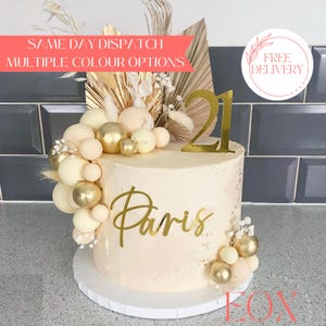 May include: A beige cake decorated with gold and white round decorations, a gold number 21 topper, and the word "Paris" in gold script. The cake is on a white cake stand and has a floral arrangement with dried palm leaves on top.
