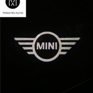 May include: A white Mini Cooper logo with wings on a black background. The logo is illuminated and appears to be a projected image.