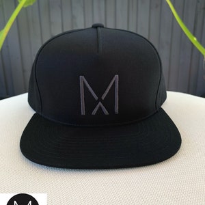 Pode incluir: Um bon&eacute; de beisebol preto e cinza com um grande "M" bordado na frente. O bon&eacute; tem uma aba plana e uma coroa curva.
