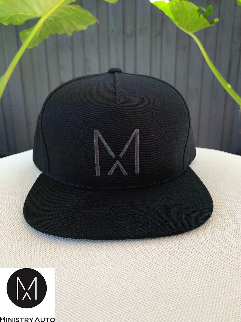 Pode incluir: Um bon&eacute; de beisebol preto e cinza com um grande "M" bordado na frente. O bon&eacute; tem uma aba plana e uma coroa curva.