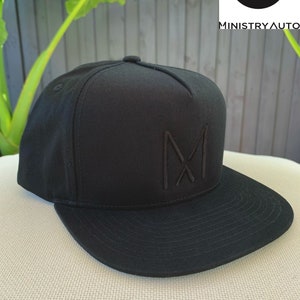 Pode incluir: Um bon&eacute; de beisebol preto com um logotipo "M" bordado em preto na frente. O logotipo &eacute; um "M" estilizado com duas linhas diagonais que se cruzam no centro.