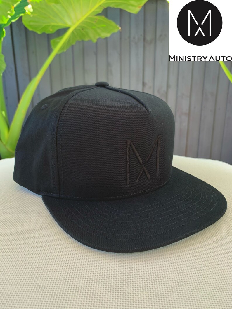 Pode incluir: Um bon&eacute; de beisebol preto com um logotipo "M" bordado em preto na frente. O logotipo &eacute; um "M" estilizado com duas linhas diagonais que se cruzam no centro.