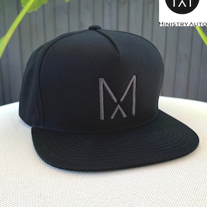 Pode incluir: Bon&eacute; de beisebol preto e cinza com um logotipo "M" bordado em cinza na frente. O bon&eacute; tem uma aba plana e uma coroa estruturada.