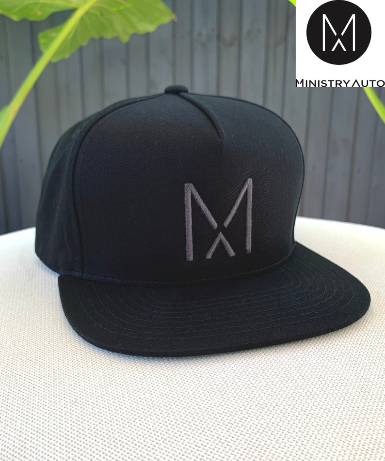 Pode incluir: Bon&eacute; de beisebol preto e cinza com um logotipo "M" bordado em cinza na frente. O bon&eacute; tem uma aba plana e uma coroa estruturada.