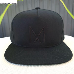 Pode incluir: Um bon&eacute; de beisebol preto de aba plana com um "M" bordado em preto na frente.