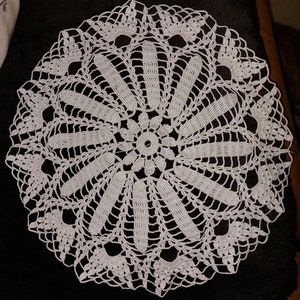 Könnte beinhalten: Weiße Häkeldeckchen mit einem zarten Blumenmuster. Das Deckchen ist rund und hat eine Mitte mit einer kleineren Blume.