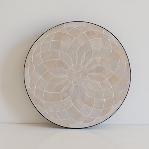 May include: A round decorative tray with a mosaic design. The tray features a pattern of small, light-colored tiles arranged in a circular, geometric design. The tiles are set against a black border.