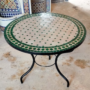 May include: A round mosaic tile table with a black metal frame and legs. The tabletop features a pattern of white and green tiles. The table is approximately 30 inches in diameter.