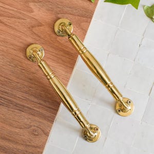 Könnte beinhalten: Zwei polierte Messing-Schrankgriffe mit klassischem Design. Jeder Griff hat eine abgerundete Basis, einen zylindrischen Griff und dekorative Details. Die Griffe werden auf einer Holzoberfläche und einer weißen Fliesenoberfläche gezeigt.