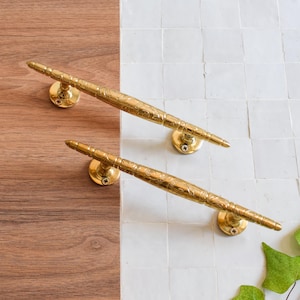 May include: Two ornate brass door pulls with floral detailing. Each pull has a long, cylindrical handle and is mounted on a round base. The pulls are set against a backdrop of wood and white tiles.