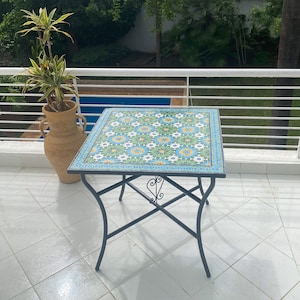 Op de afbeelding: Een vierkante, betegelde tafel met een geometrisch patroon in groen, blauw en geel. De tafel heeft een zwart metalen frame met een decoratief scroll-ontwerp.