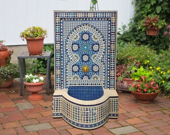 May include: A blue and white mosaic tile fountain with a traditional Moroccan design. The fountain has a curved basin and a decorative archway. The fountain is made of ceramic tile and is suitable for outdoor use.