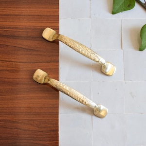 Puede incluir: Dos tiradores de latón para armarios con un patrón texturizado en forma de hoja sobre una superficie de baldosas marrones y blancas. Los tiradores tienen forma rectangular con extremos redondeados.
