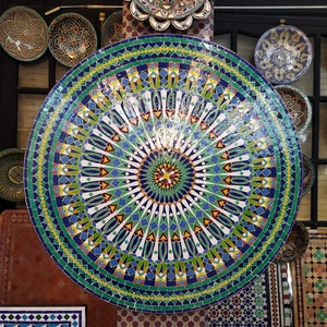 Könnte beinhalten: Eine große runde Tischplatte mit einem farbenfrohen Mosaikdesign. Das Mosaik zeigt ein zentrales Sternburst-Muster mit radial verlaufenden Linien aus grünen, blauen, gelben und roten Fliesen.