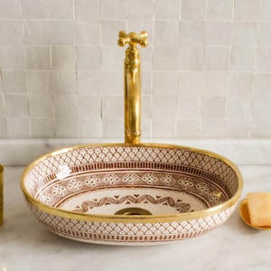 Può includere: Lavabo ovale in ceramica con un motivo geometrico marrone e bianco e un bordo dorato. Il lavabo ha un rubinetto dorato con una maniglia a forma di croce.