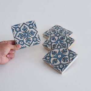 Può includere: Sottobicchieri quadrati in ceramica con un motivo floreale blu e bianco. Un sottobicchiere è tenuto in mano, mentre altri tre sono disposti nelle vicinanze. I sottobicchieri hanno un motivo decorativo e uno sfondo chiaro.