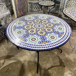 May include: Round mosaic table with a blue, white, and gold geometric pattern. The table has a black metal base and is approximately 36 inches in diameter.