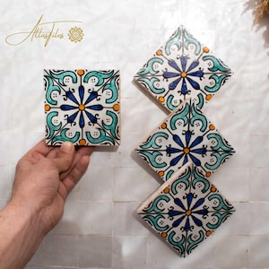 May include: Hand-painted ceramic tiles with a floral design in blue, teal, and orange. One square tile is held by a hand, while three diamond-shaped tiles are arranged on a white tiled surface. The tiles feature a central flower motif.