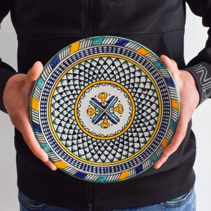 May include: A decorative ceramic plate with a hand-painted design. The plate features a central motif with yellow, blue, and turquoise accents, surrounded by a geometric pattern. The rim has a repeating pattern of blue, yellow, and turquoise. The plate is held by a person.